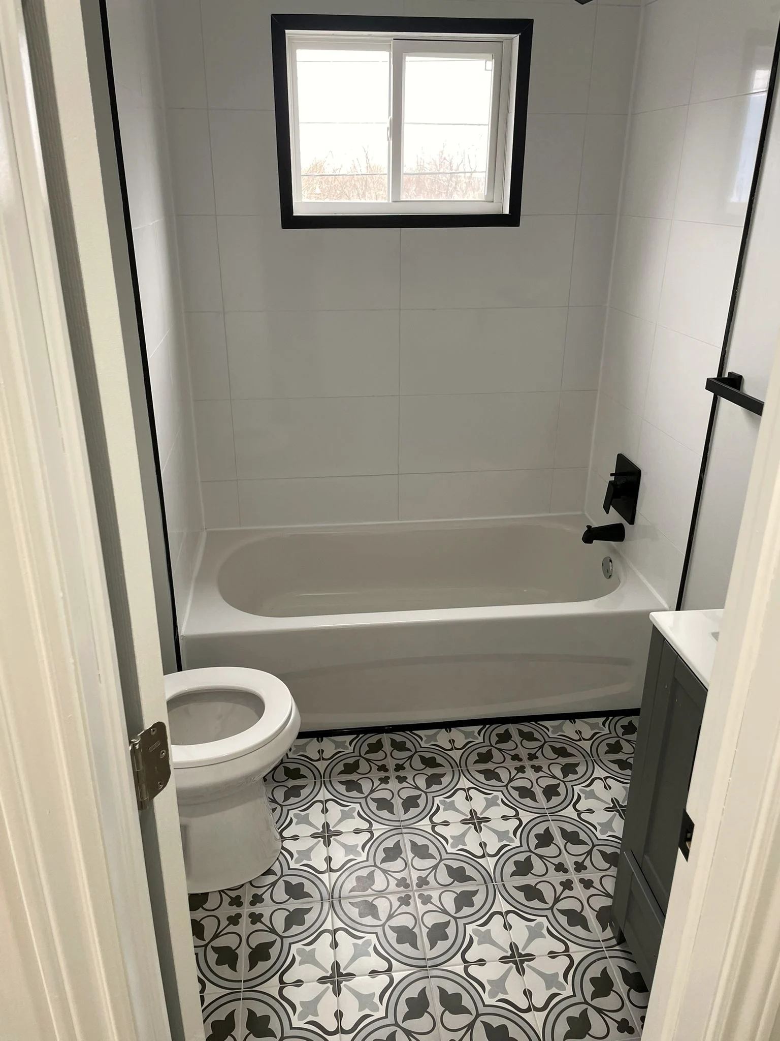 Finished bathroom with a white tub under a black framed window, large format white wall tile, and black and white patterned floor tile.