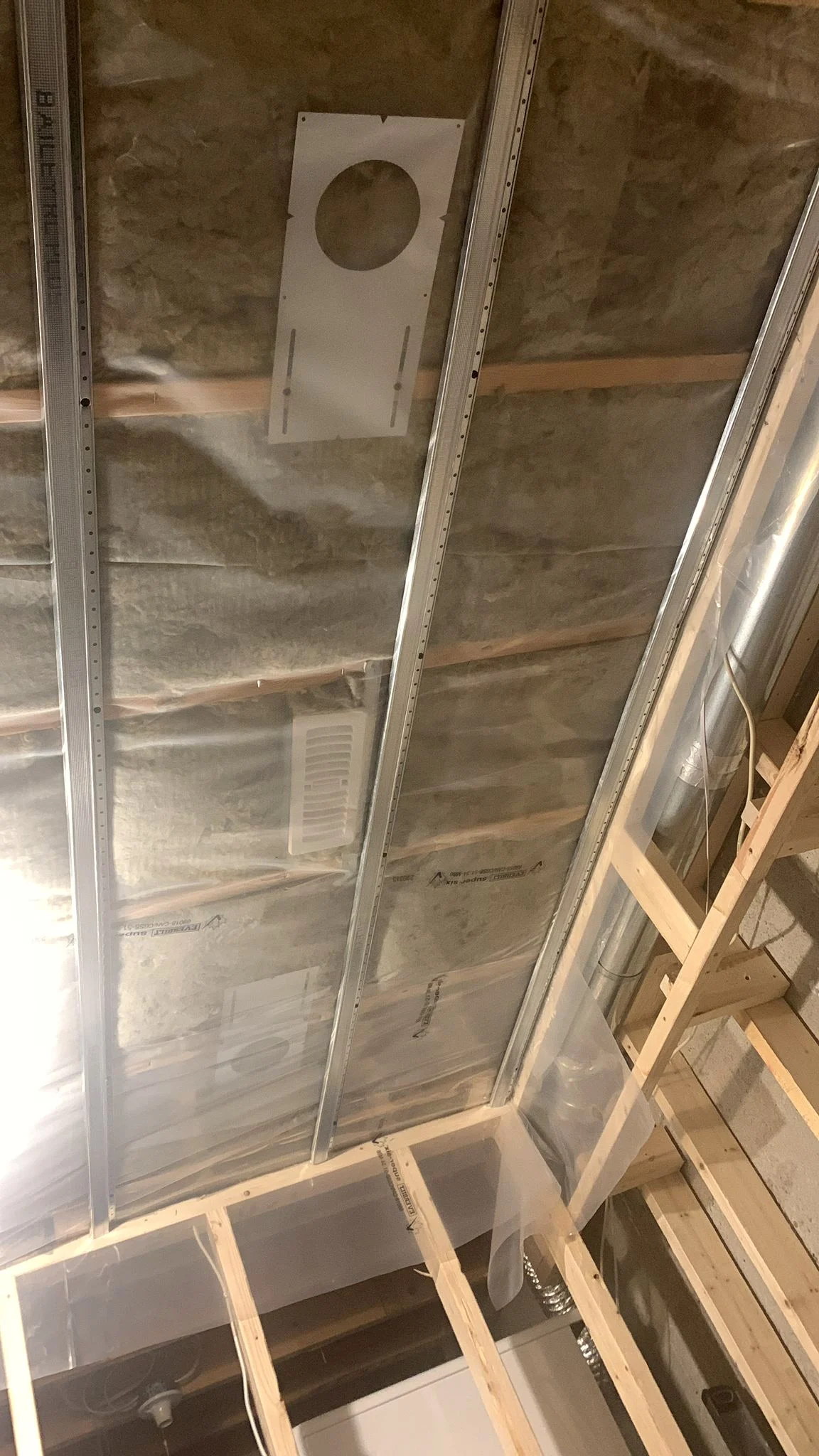 Ceiling insulation between the joists with metal channel screwed across them, a round duct overhead, and a white register set into the framing.