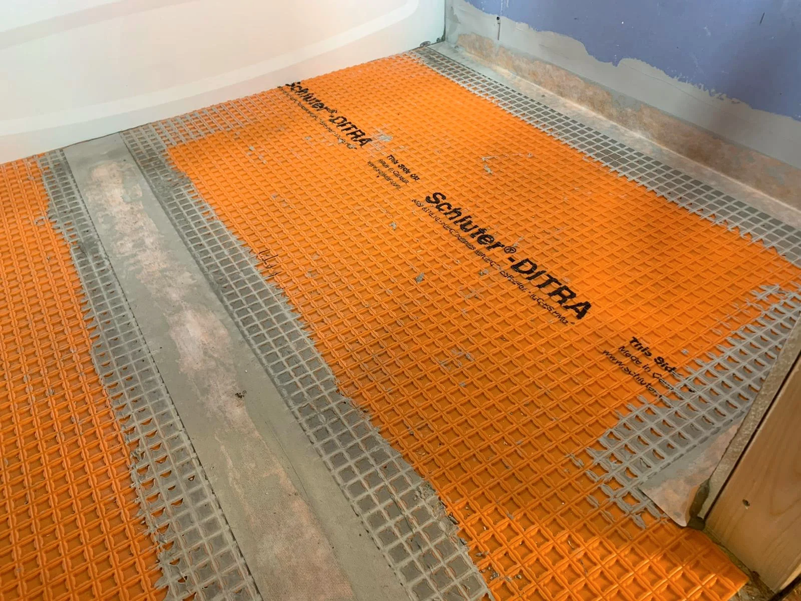 Schluter DITRA membrane laid on the floor beside a curved shower base, with blue painted drywall behind.