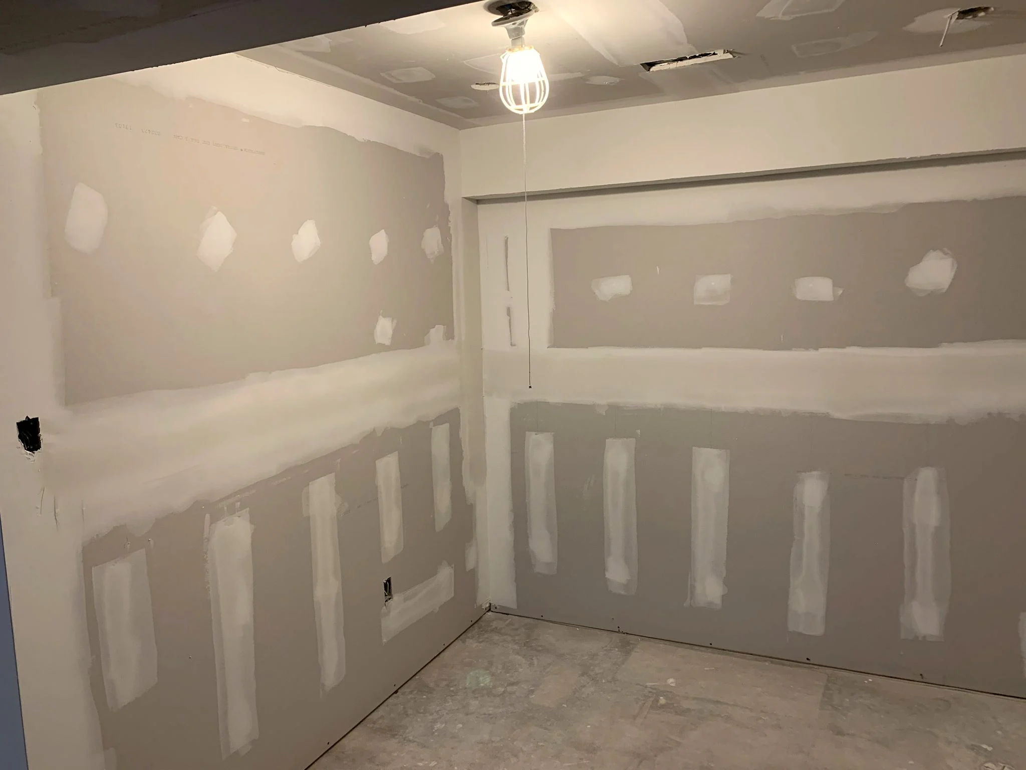 Basement room mid-renovation with taped and mudded drywall and a bare bulb hanging from the ceiling.