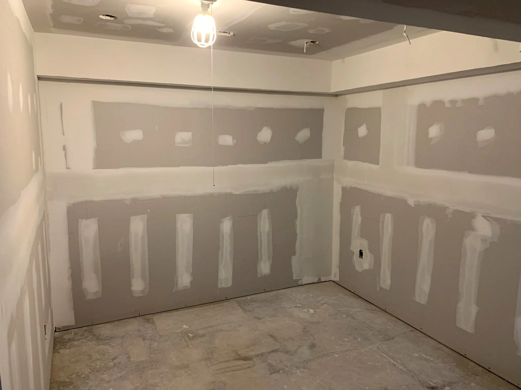 Basement room mid-renovation with taped drywall, recessed light cutouts in the ceiling, and a caged work light hanging over the bare concrete floor.