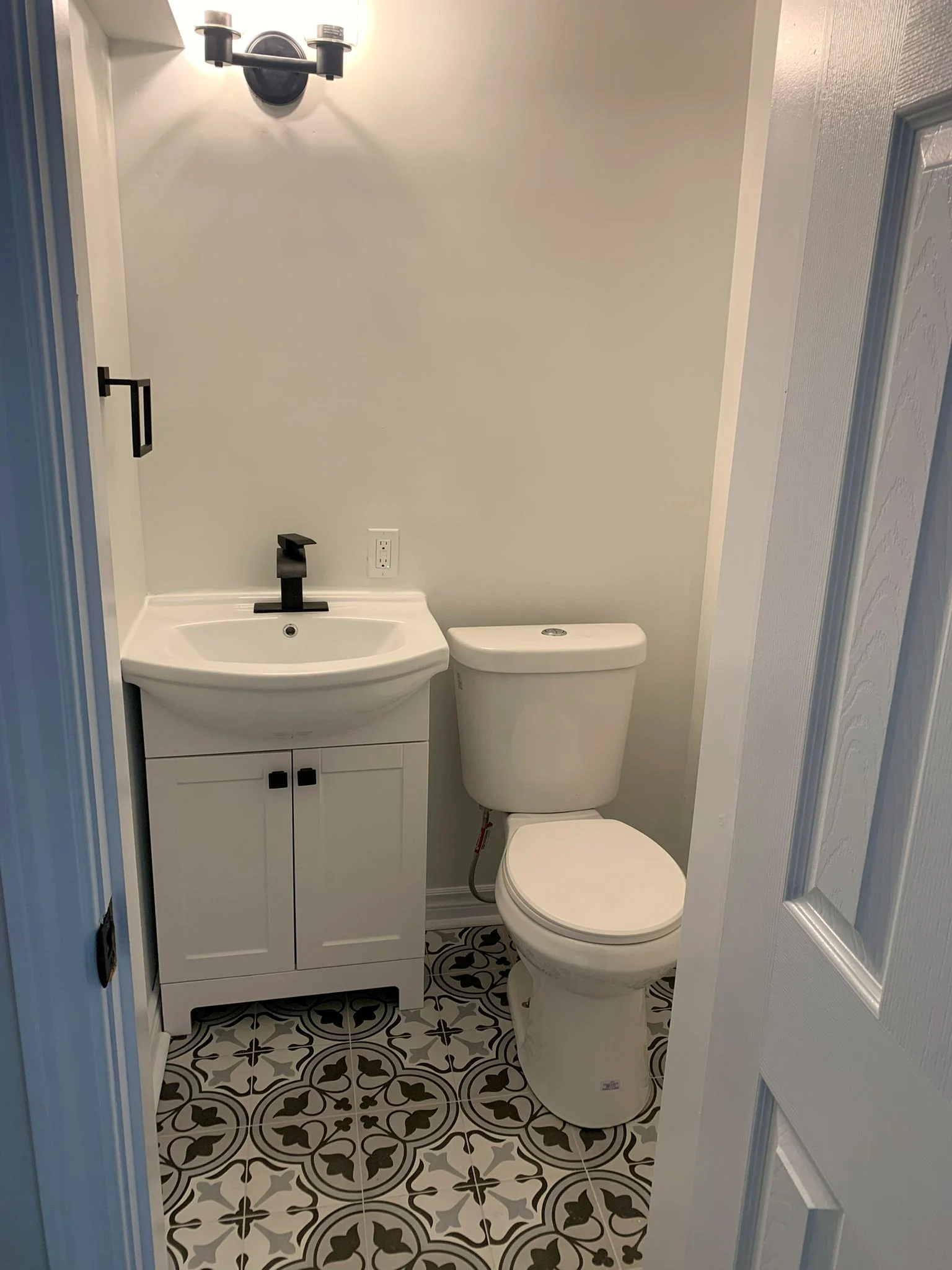 Finished powder room with black and white patterned floor tile, a white two door vanity with a black faucet, and a black sconce above.
