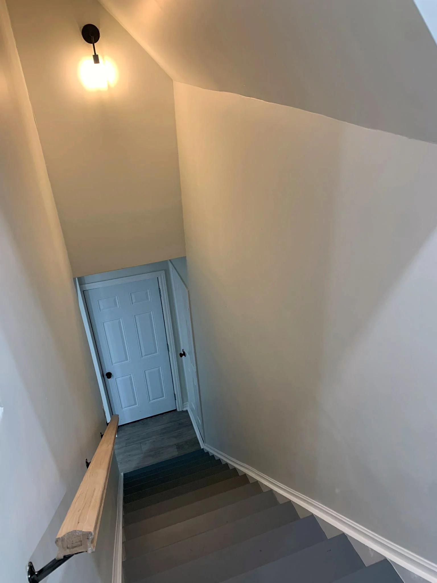 Staircase looking down to a white panelled door at the bottom, with a wood handrail, grey treads and white risers, and a black sconce on the wall.