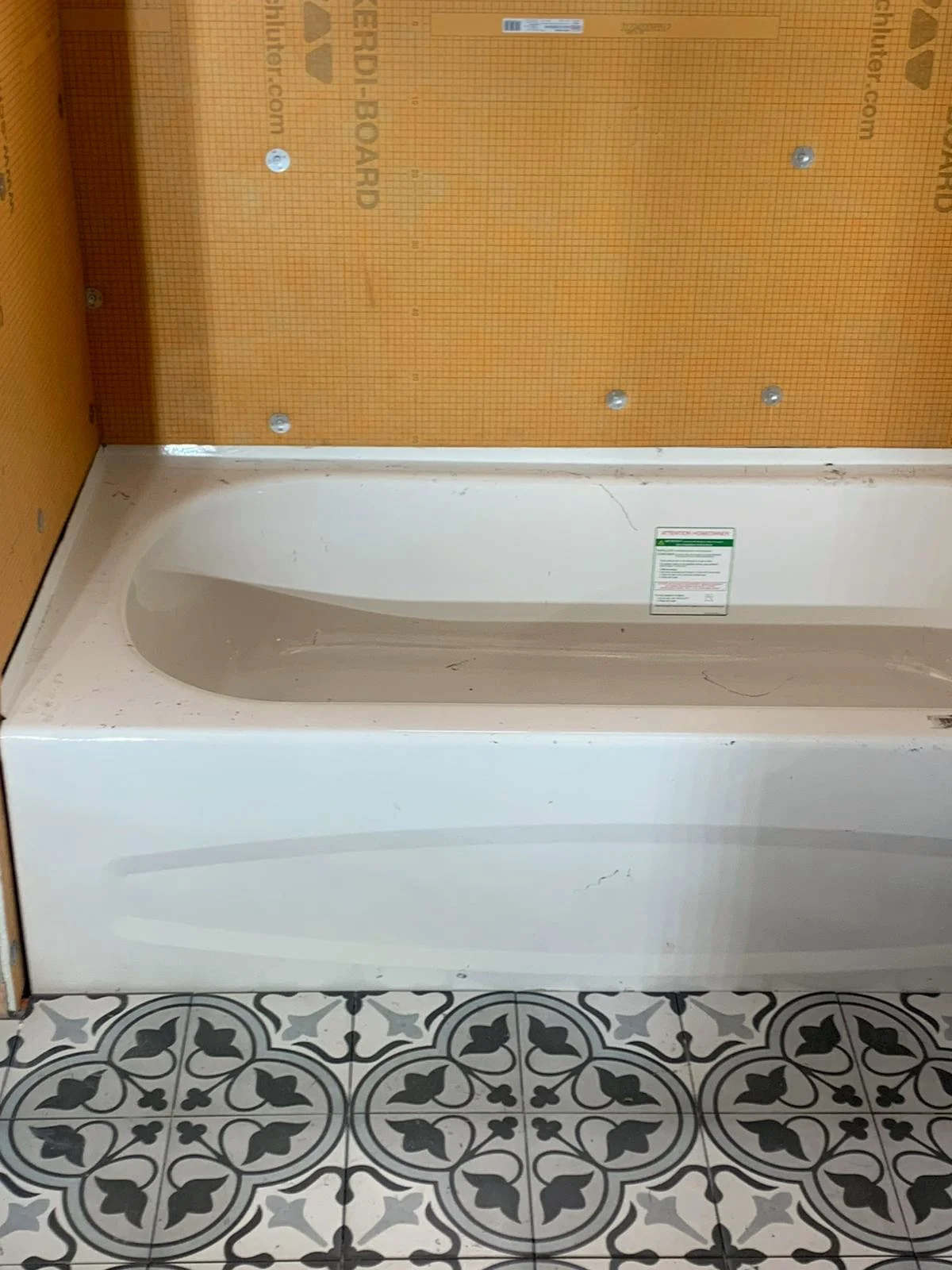 Bathtub installed against an orange Kerdi board wall, with black and white patterned floor tile already laid.