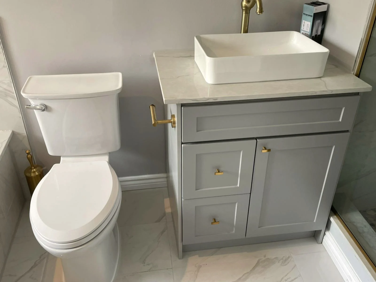 Finished bathroom vanity with a grey shaker cabinet, a white vessel sink, and a brushed gold faucet on marble-look tile.