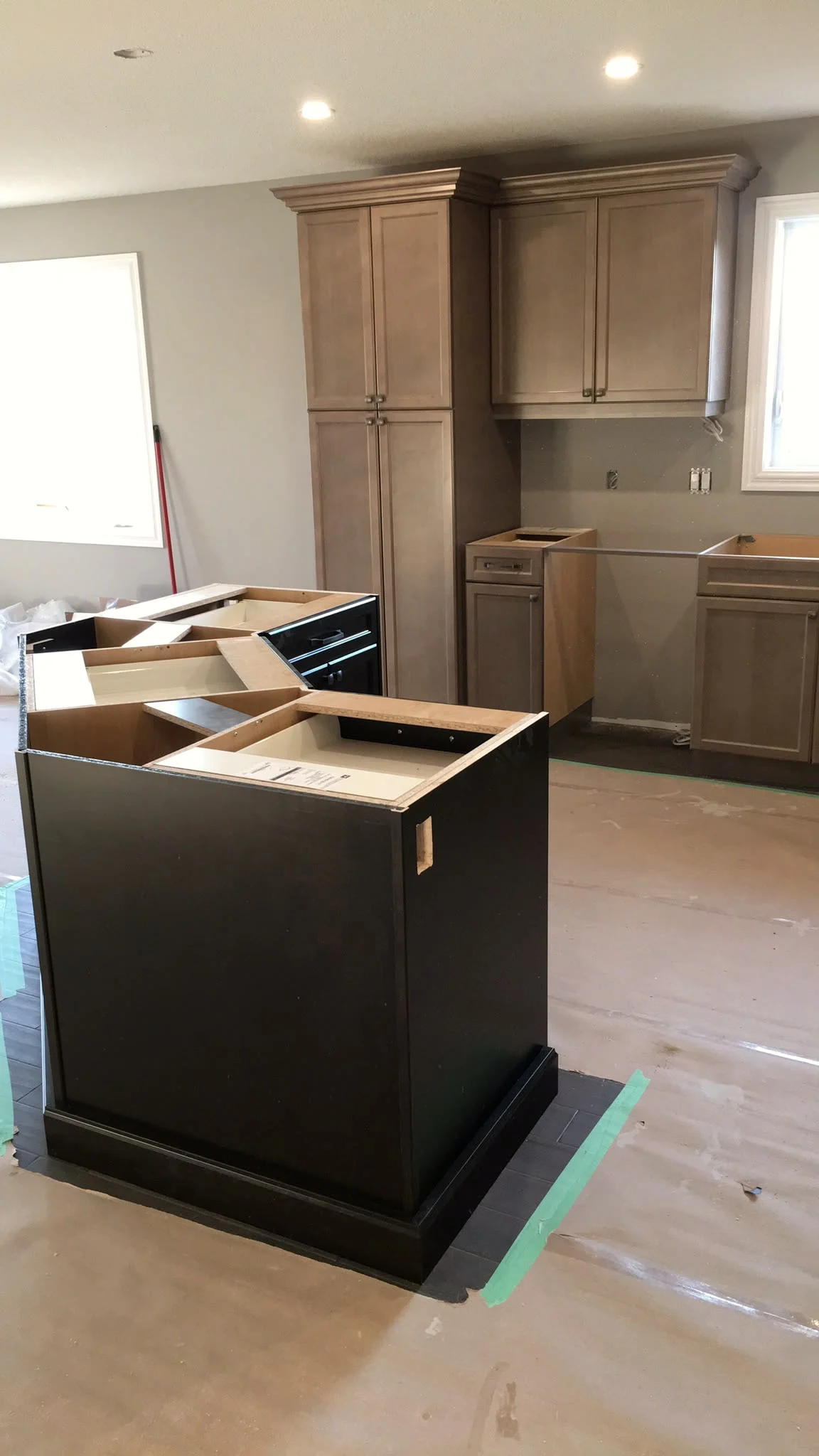 Kitchen mid-install with tan cabinetry along the walls, a dark espresso island base still open at the top, and the floor covered in protective paper.