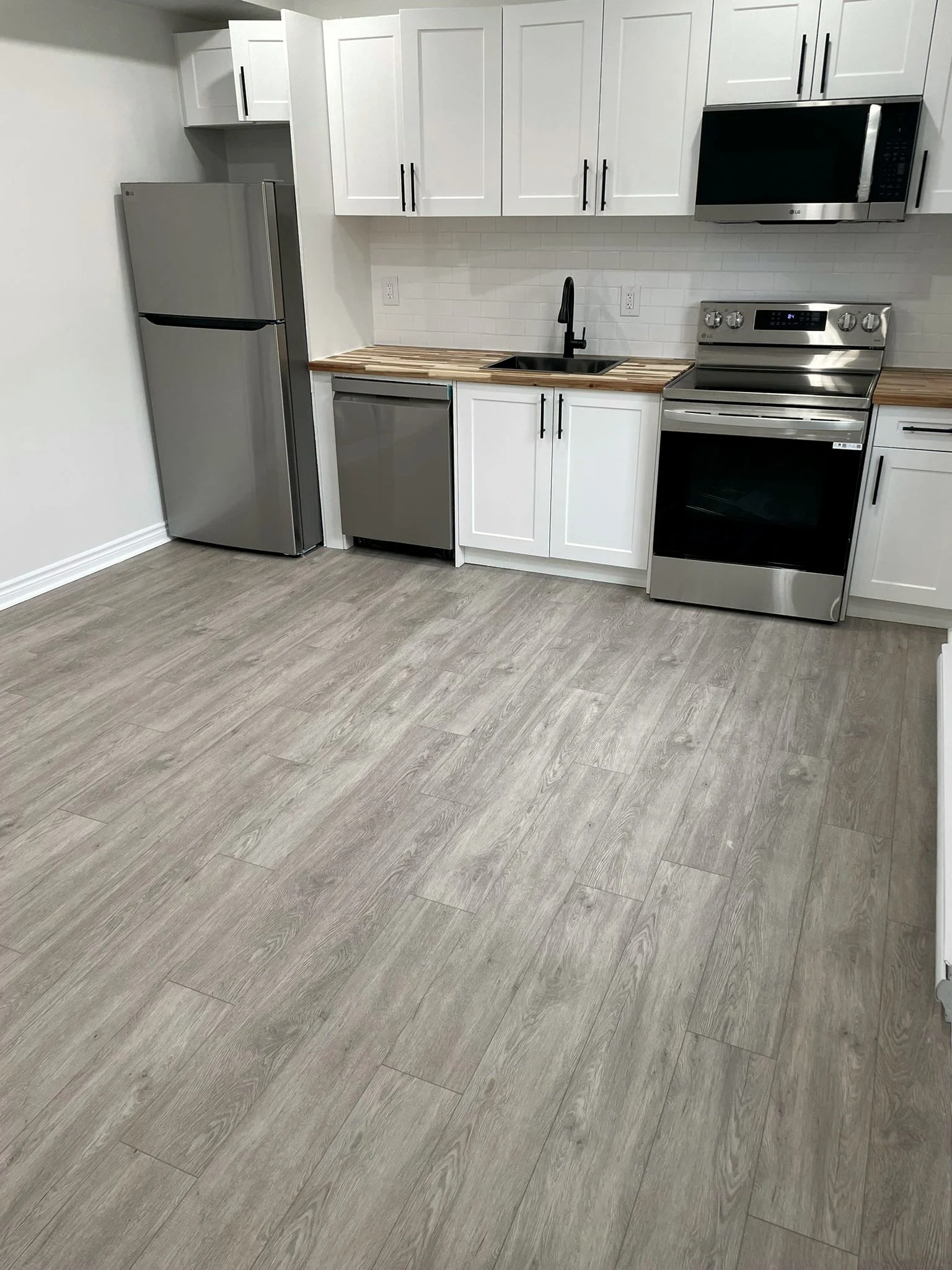 Finished kitchen with white shaker cabinets, a butcher block counter, and stainless steel appliances on a grey plank floor.