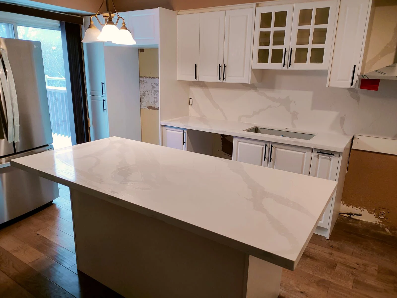 Kitchen mid-renovation with white cabinets, a marble-look stone island top and matching slab backsplash installed, and an empty sink cutout beside a wall still torn open.
