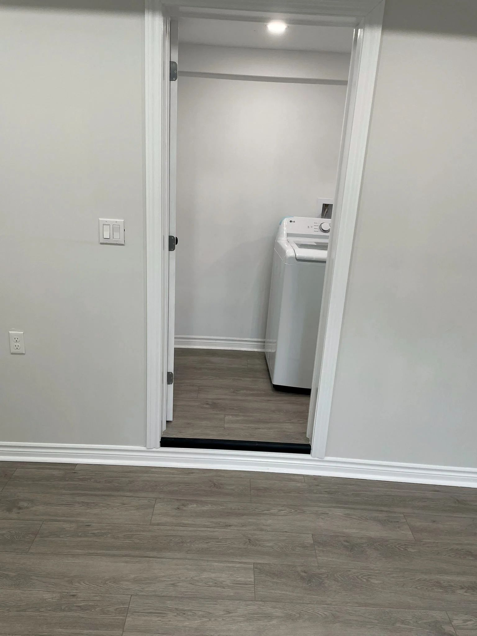 Finished laundry closet with a white top load washer, seen through the open door from a room with grey plank flooring.