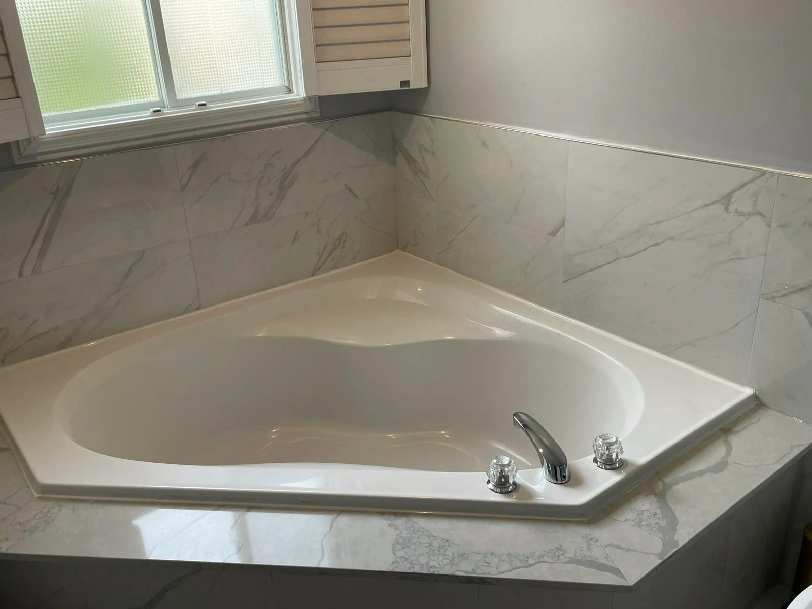 Finished corner soaker tub set into a marble-look tiled deck, with a shuttered window above it.