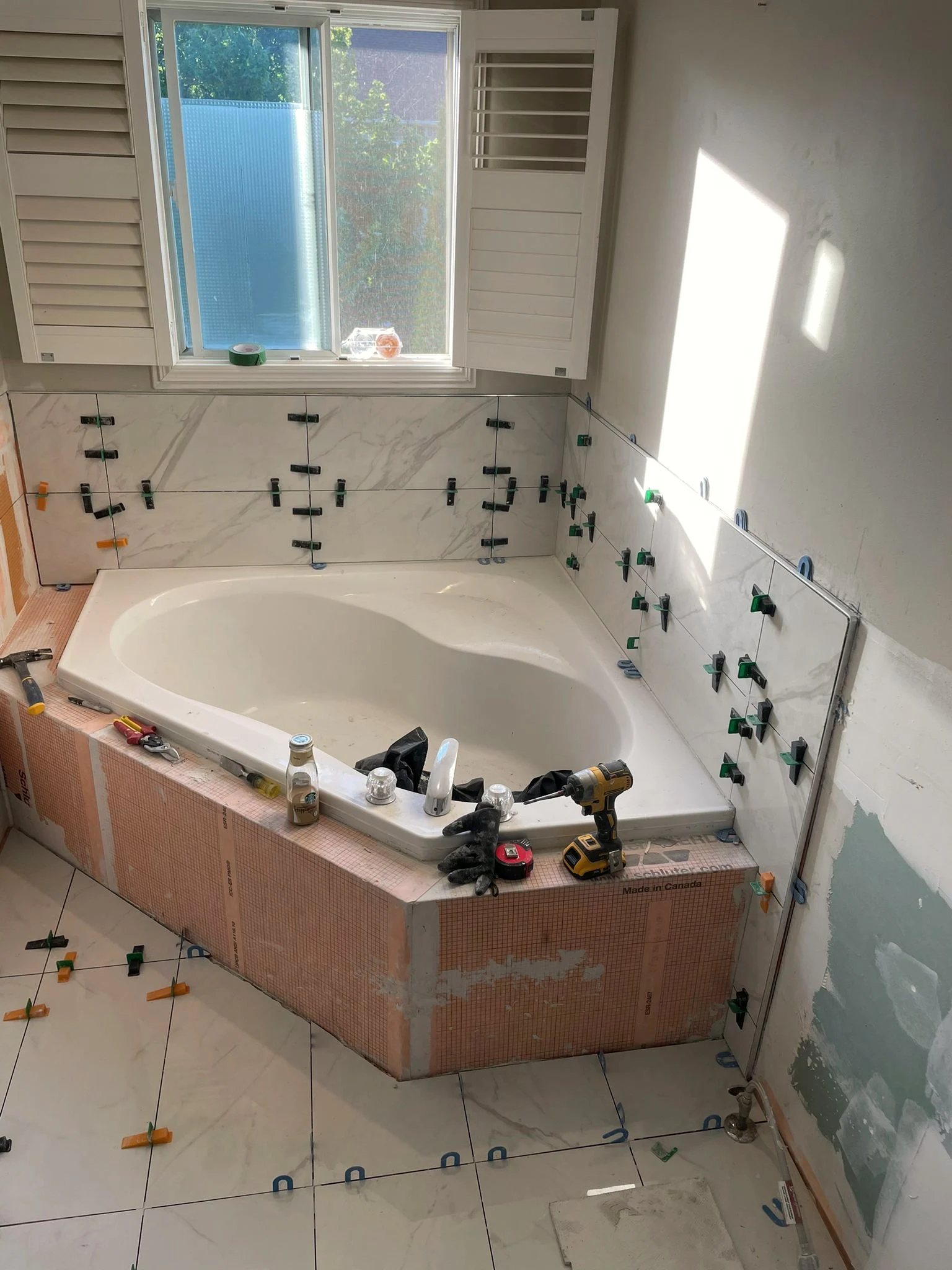 Corner soaker tub mid-build, with large format marble-look tile held on leveling clips and the tub deck still wrapped in orange Kerdi board.