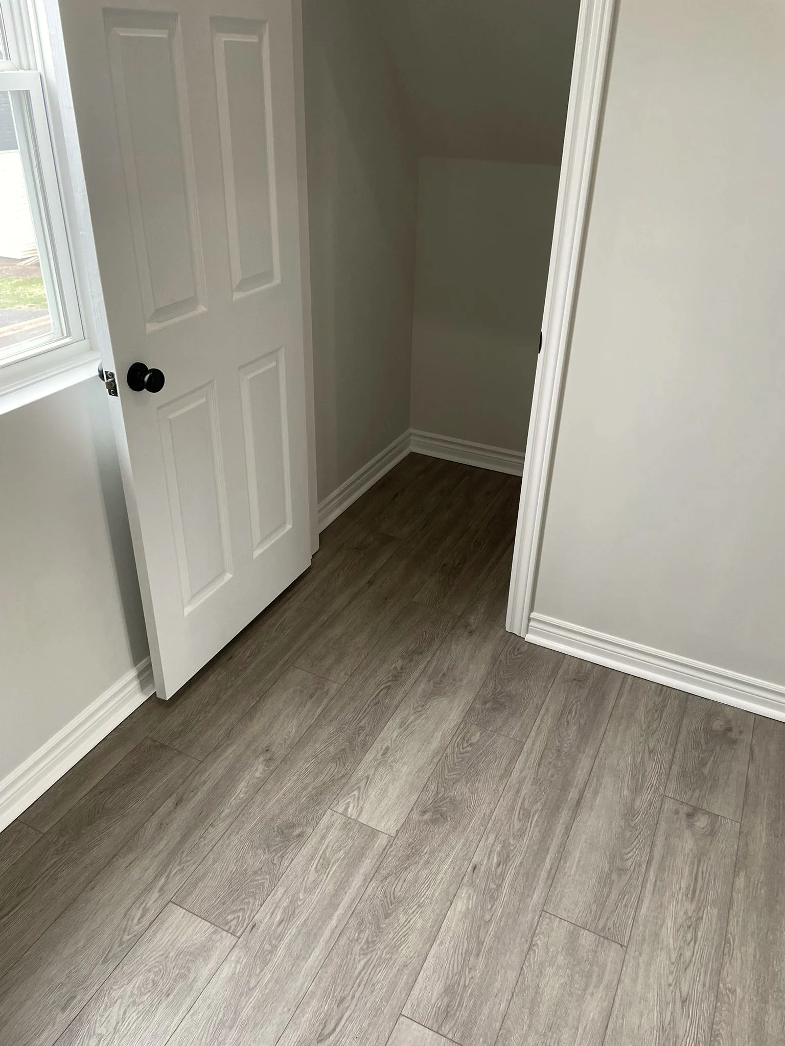 Open white panelled door revealing a small storage nook with a sloped ceiling, with grey plank flooring running through.