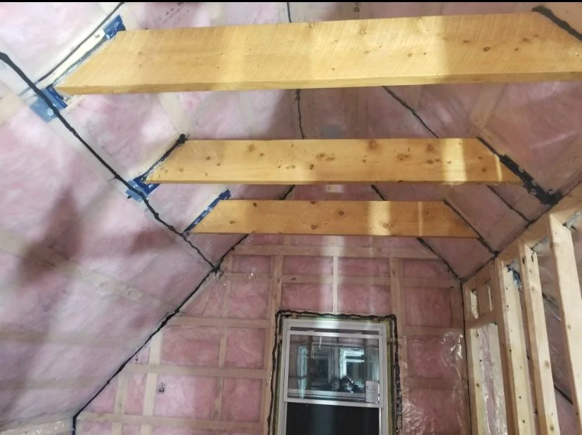 Pitched roof rafters with pink fibreglass batt insulation behind a taped vapour barrier, with a window in the end wall below.