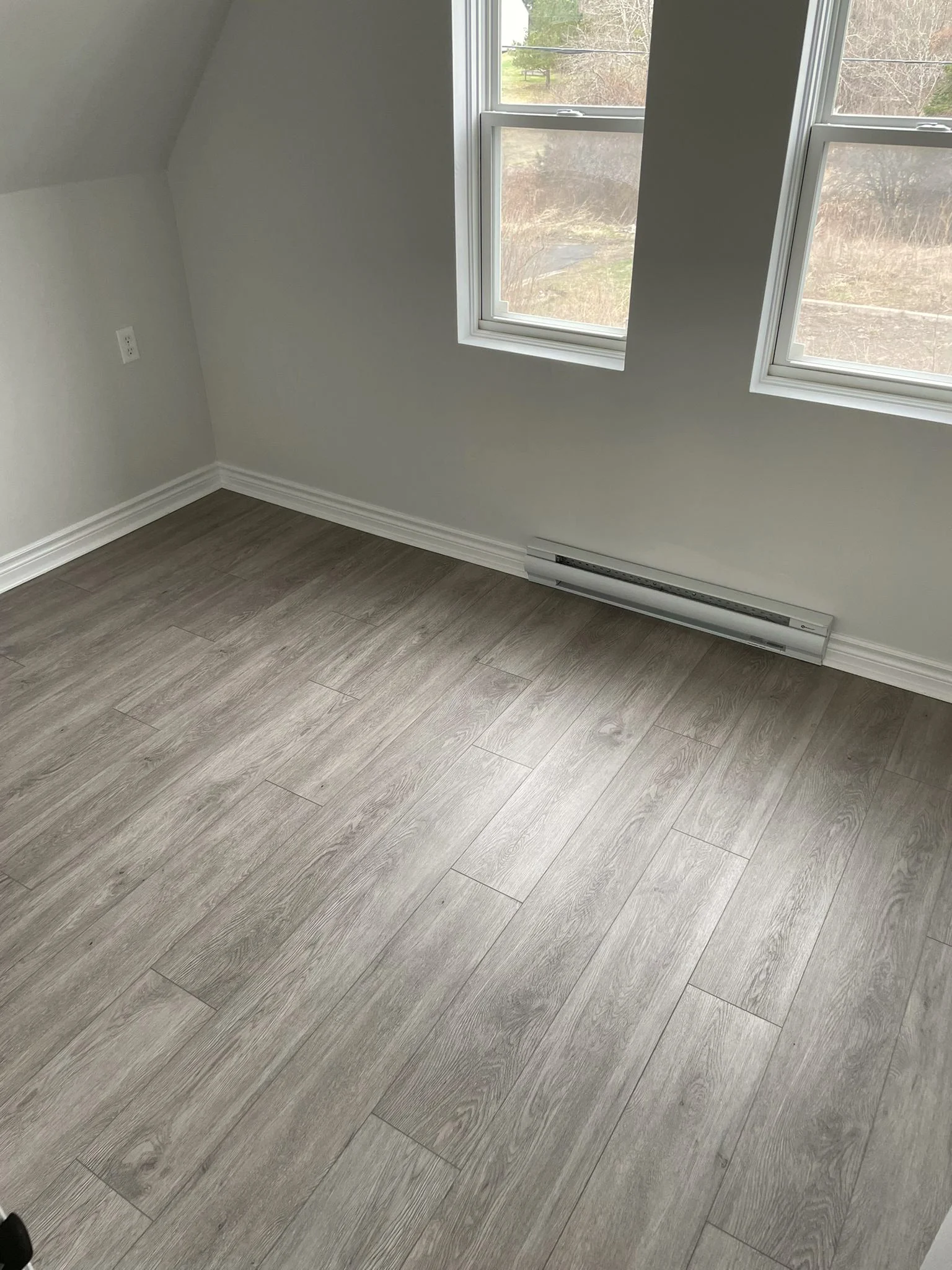 Empty room with a sloped ceiling, two windows looking out over the yard, an electric baseboard heater, and grey plank flooring.